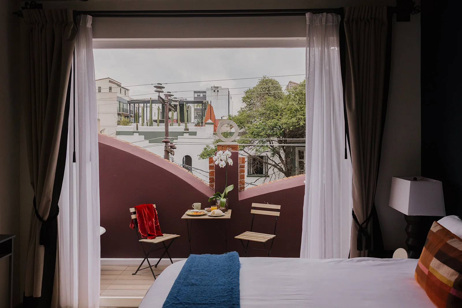 The private balcony seen from the bed through the open doors: a bistro table laid, the curved wine-coloured wall and the Roma rooftops
