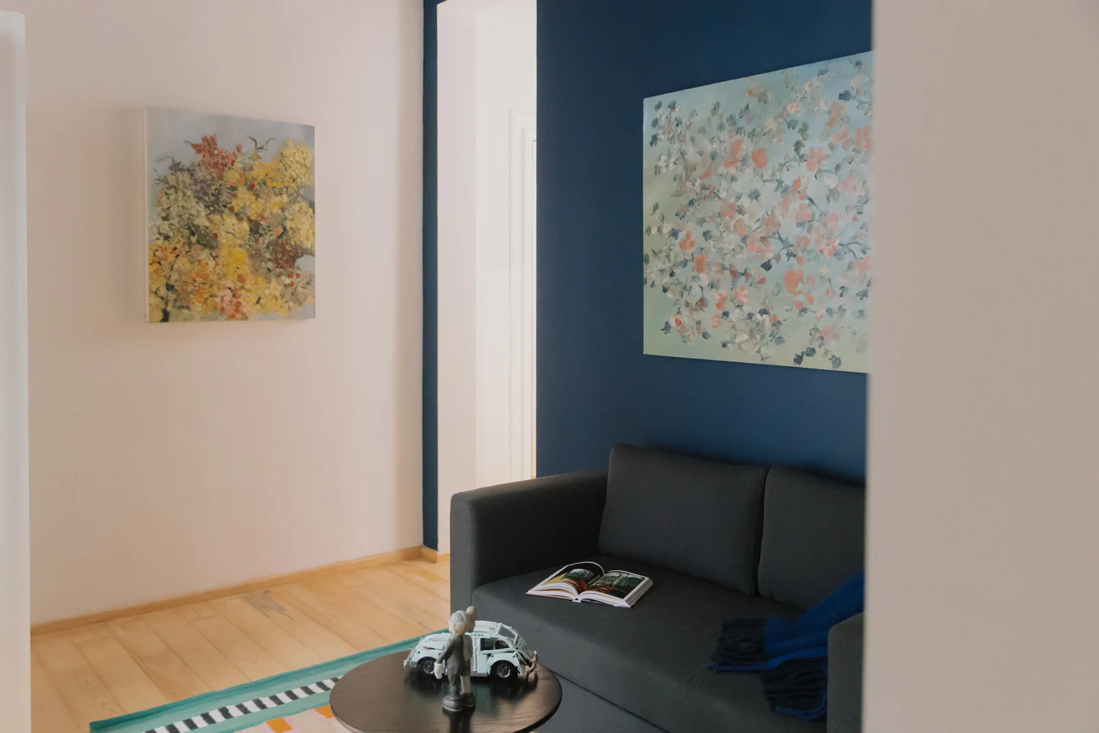 The studio's living area: a grey sofa against the blue wall under a floral painting, with a round turned-wood table