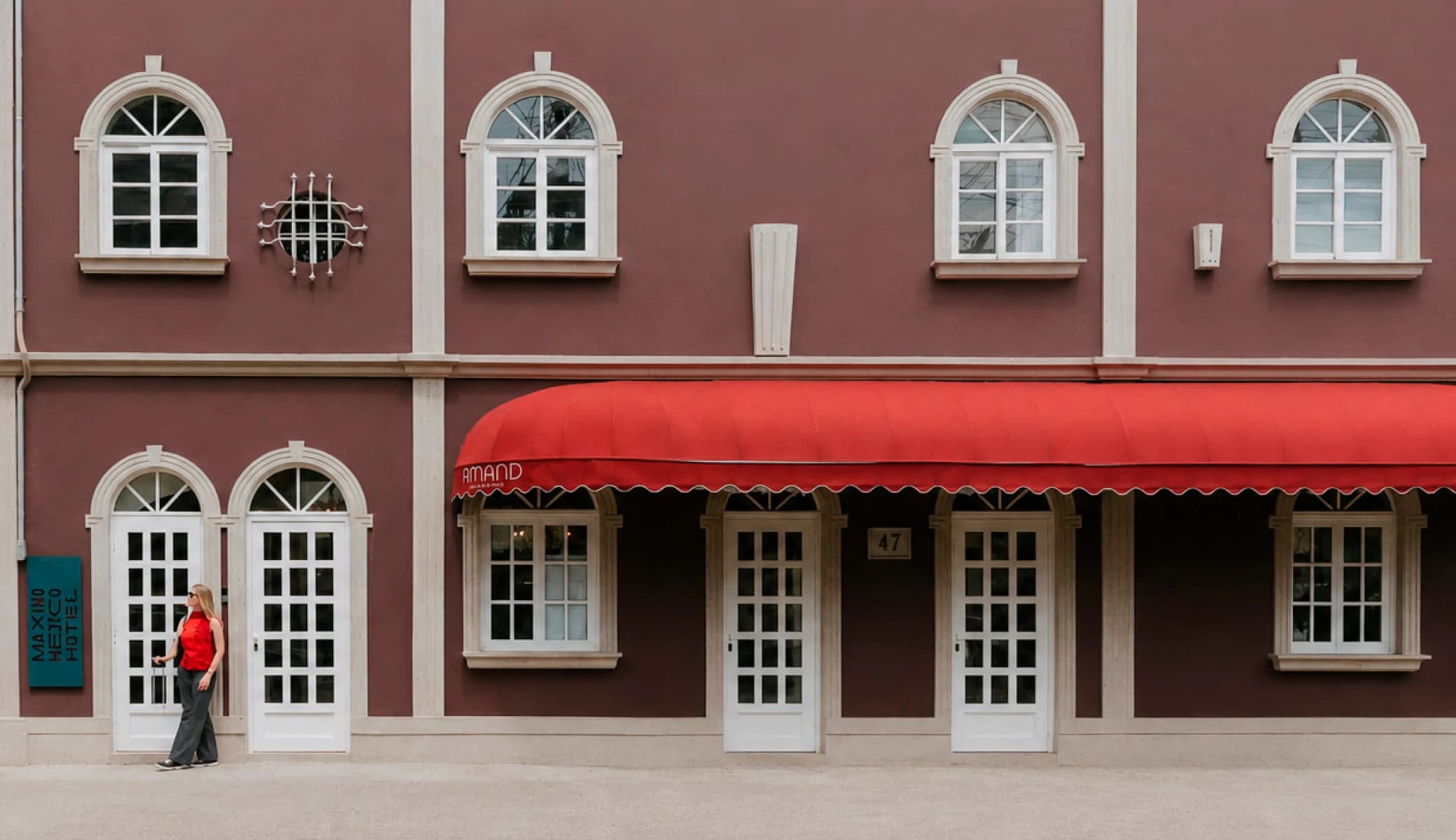 The facade of Máximo México Hotel on Calle Manzanillo: a wine-coloured wall with white arched windows, the house's turquoise vertical sign, Amand's red awning, and a guest arriving with a suitcase