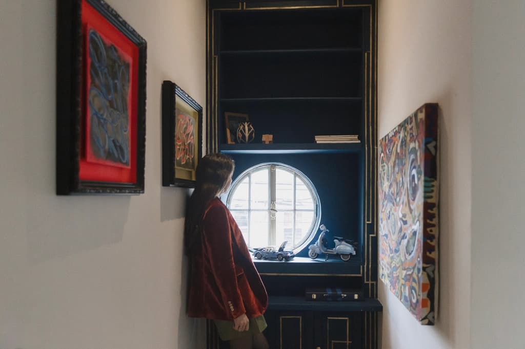 The house's entrance hall: a corridor with curated art on both walls, the porthole window at the far end, and a guest looking at the paintings