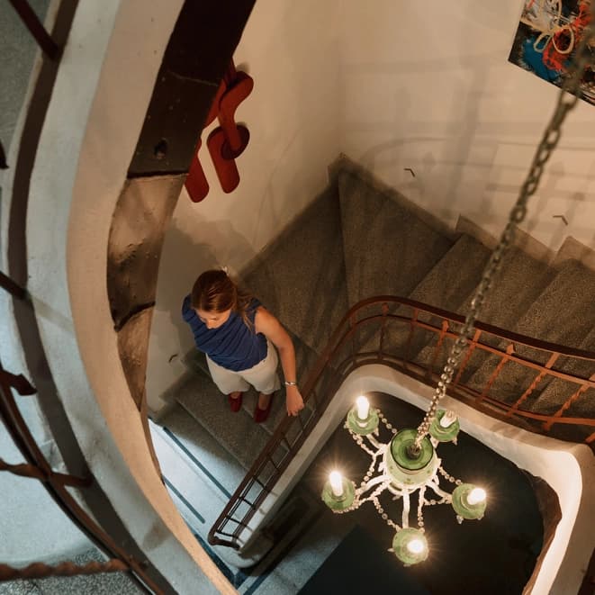The curved staircase seen from above, with a guest coming down and the green glass chandelier hanging in the well between the wrought-iron banisters