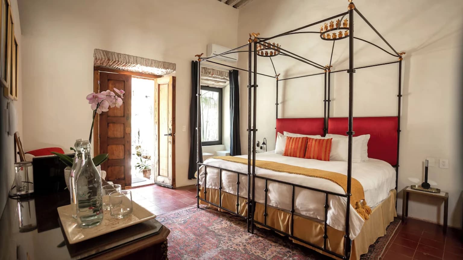 A bedroom with a wrought-iron four-poster bed and a red headboard, a Persian rug and an orchid on the table