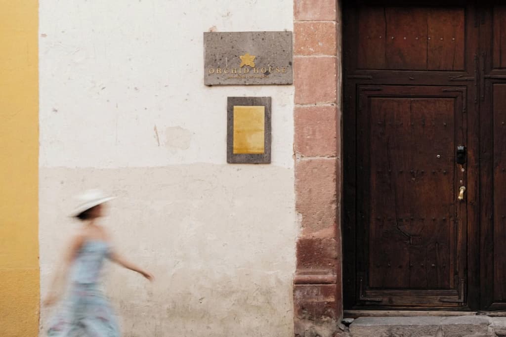 The house's facade on the street: the stone plaque with the gold orchid and the Orchid House name beside the wooden door, as a guest in a hat walks past