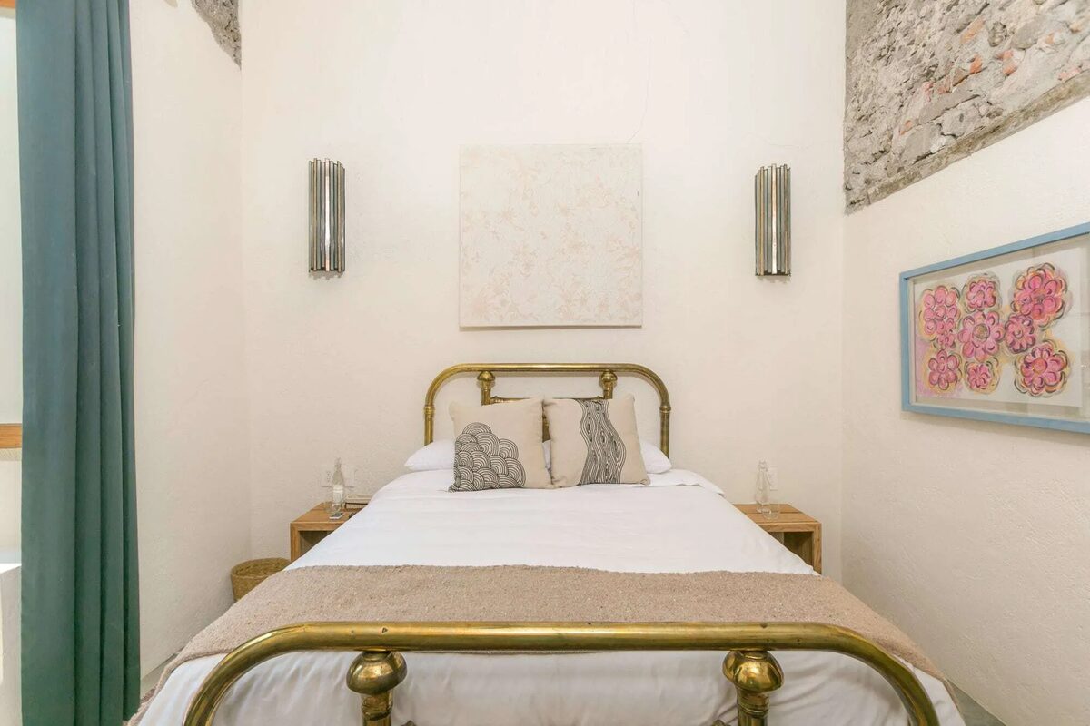 Room with a brass queen bed between two metal sconces, a stone wall in the corner and a watercolour of flowers