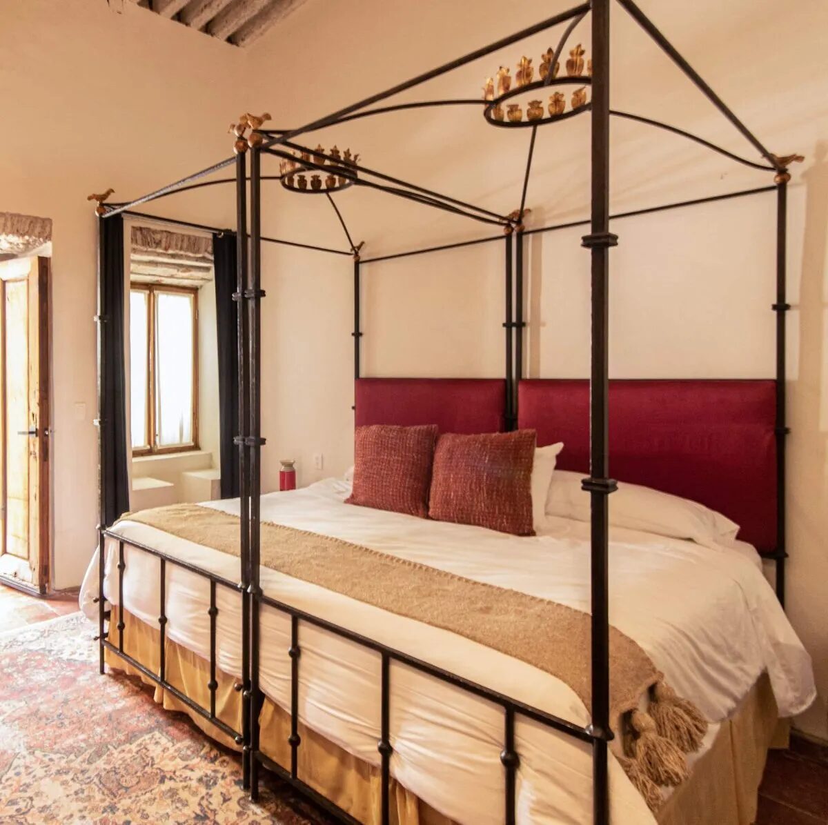 Suite with a wrought-iron four-poster king bed and a red headboard, under timber beams, beside a window with a built-in seat