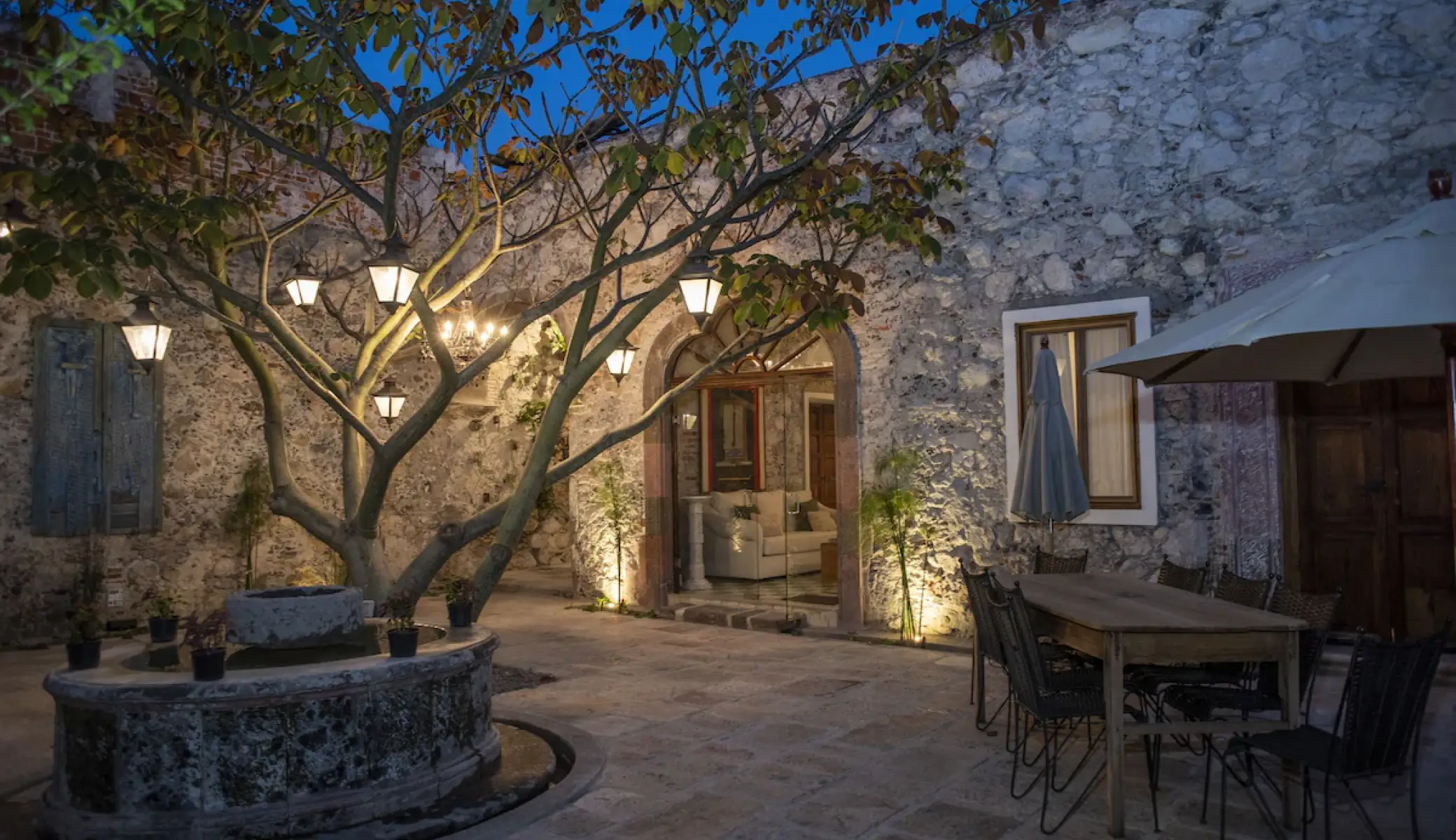 The courtyard at dusk: a tree lit with lanterns, the cantera stone fountain, and the garden table laid under a parasol