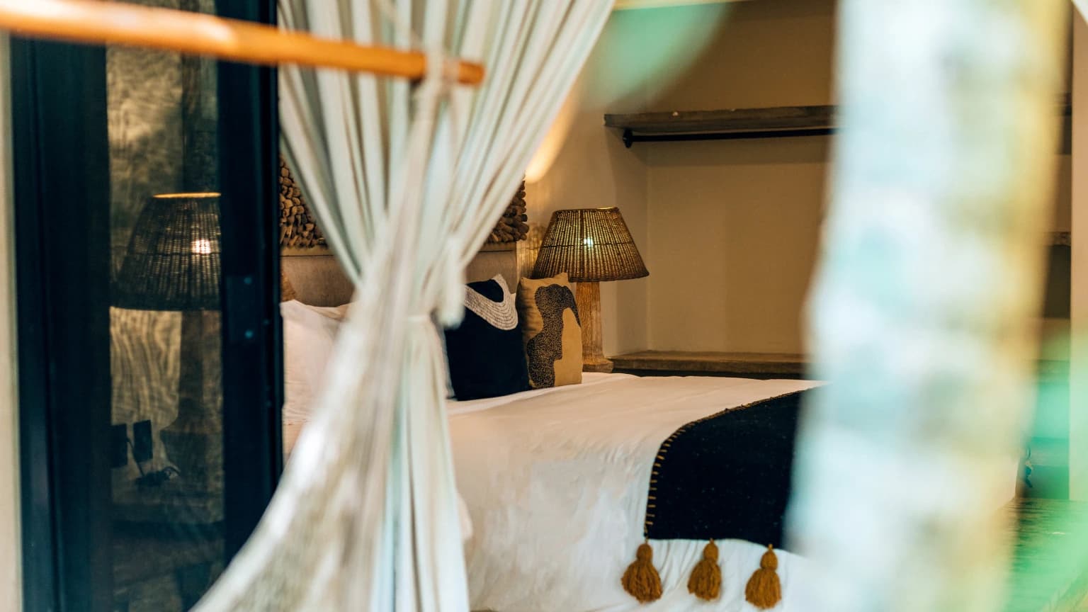 A bedroom with a canopy bed and sheer curtains, black-and-white cushions, and a wicker lamp