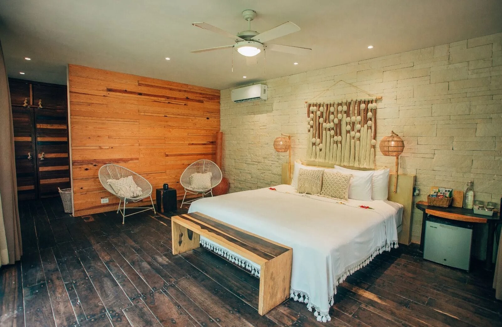 Room with a king bed beneath a macramé and tassel wall hanging, a timber-plank wall and two round wicker chairs