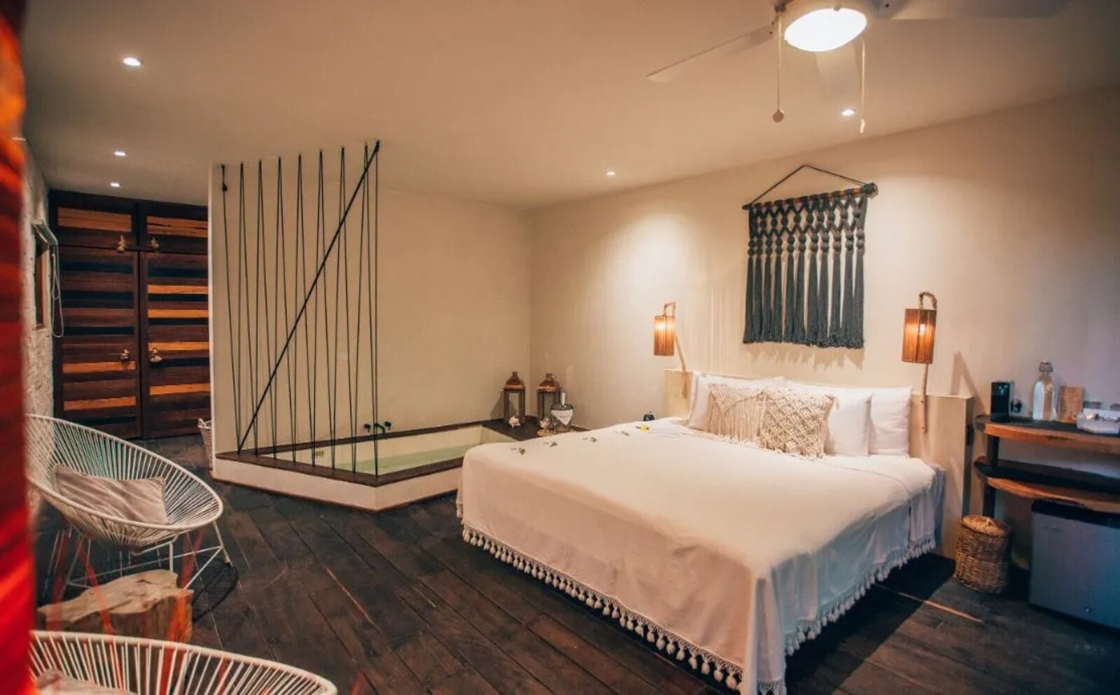 Room with a sunken tub behind a curtain of hanging cords, beside the bed with its macramé headboard