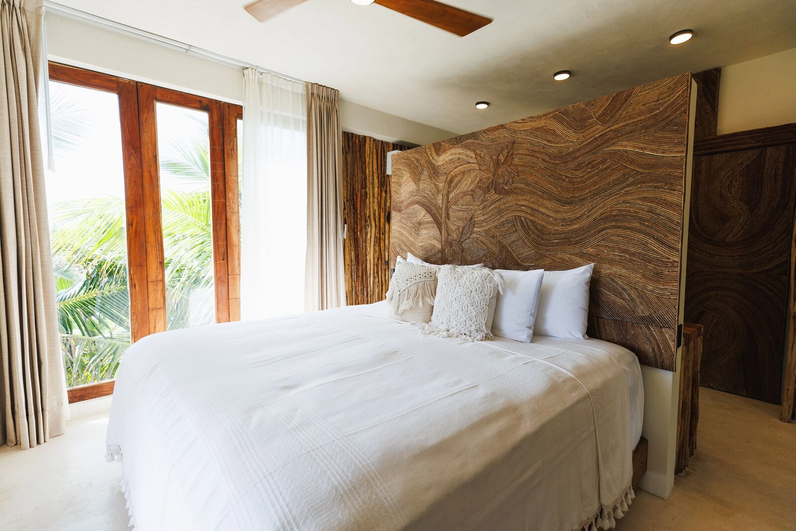 A carved timber headboard with an orchid etched into it, beside a floor-to-ceiling window open to the palm trees