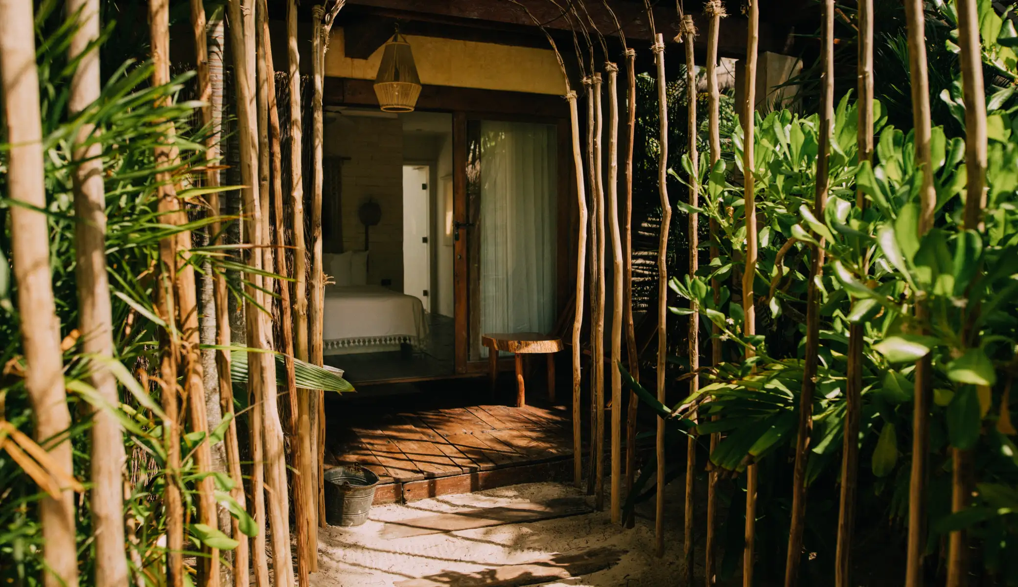 A tunnel of reed poles between palm trees leads to a room's wooden terrace, on the sand