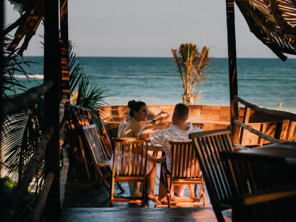 A couple toasts at a table facing the ocean, under the Orchid Beach Club's palapa