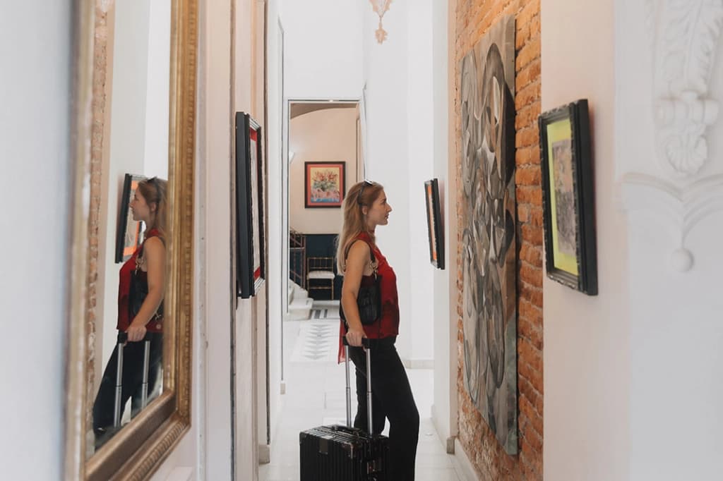 A guest with a suitcase walks down the brick corridor with framed art, reflected in a gilded mirror
