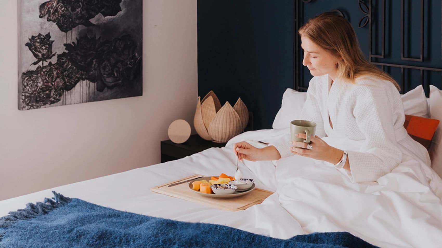 A guest in a robe has breakfast in bed, a tray of fruit and yogurt on the covers