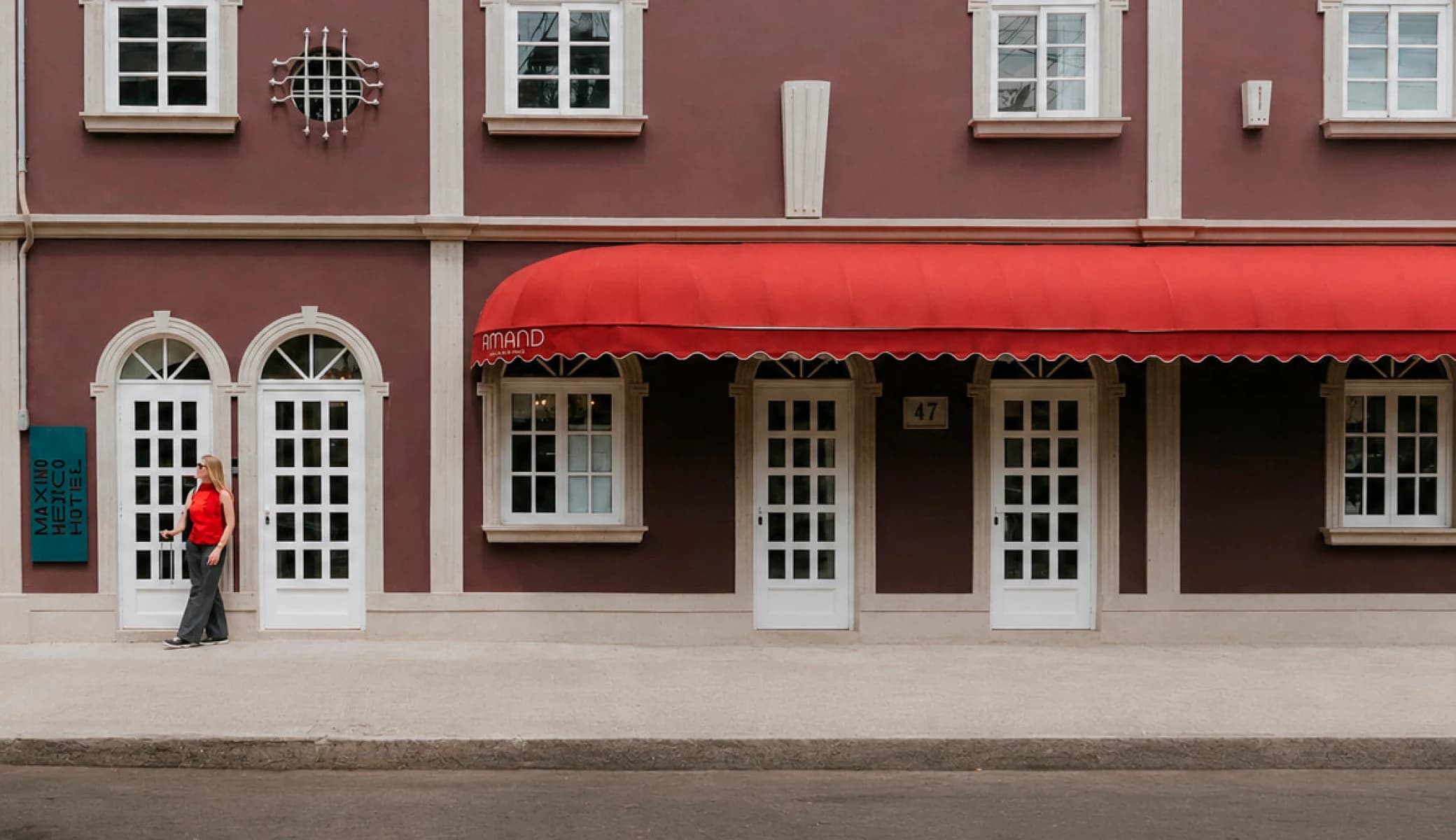 The hotel's wine-coloured facade, with the "Maximo Mexico Hotel" sign, Amand's red awning and a guest with a suitcase on the sidewalk