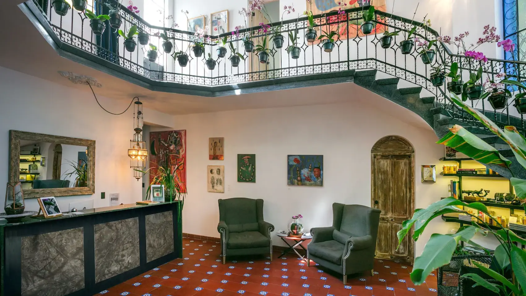 The house's lobby: the staircase with orchids hanging from its railing, green armchairs, art on the walls and the red-and-blue tiled floor