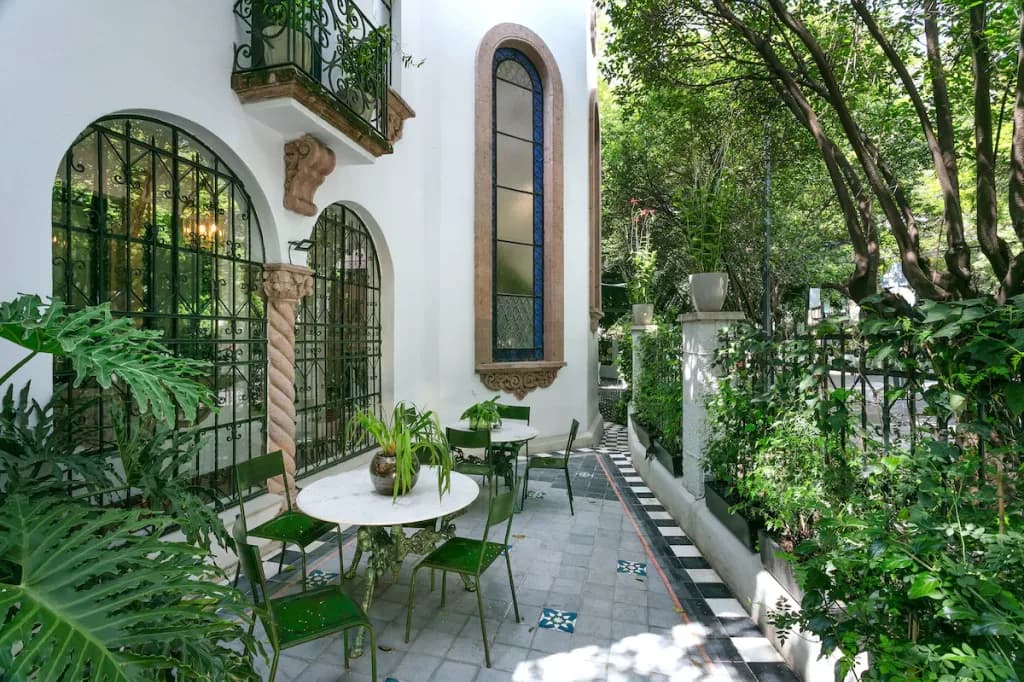 A side walkway between planters and the facade, with marble tables and green wrought-iron chairs, black-and-white tiled floor and the street gate beyond