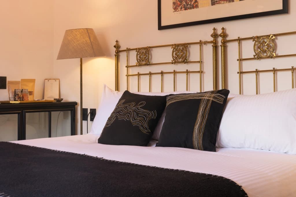 A guest room with an ornate brass headboard, white bedding, embroidered black cushions and a dark throw at the foot, beside a floor lamp and a dresser with amenities
