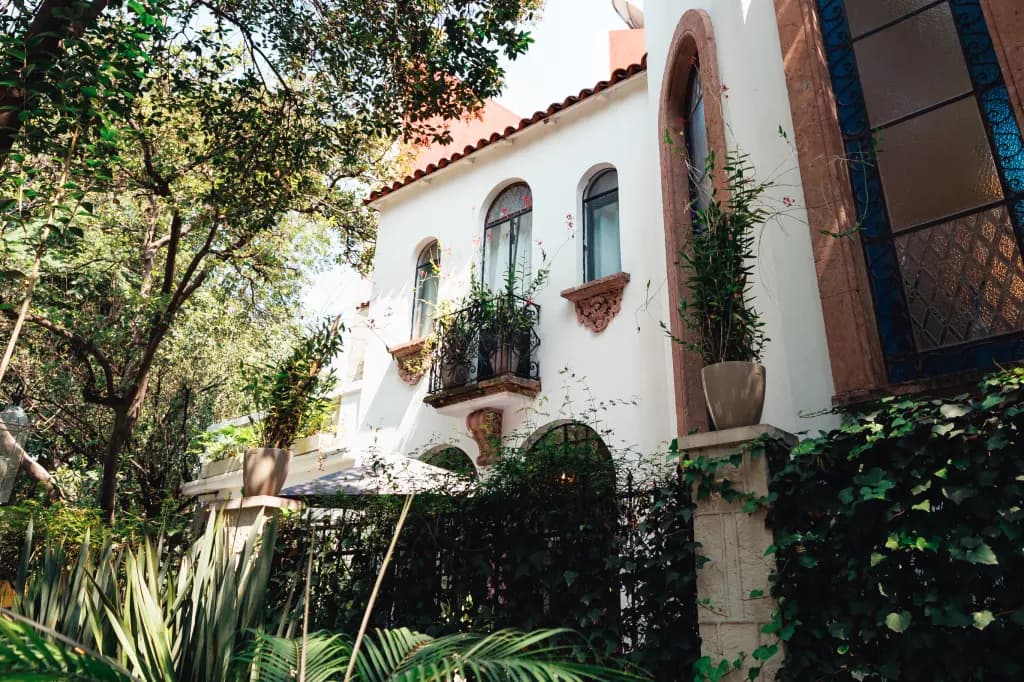 The facade seen from the garden, with its red tile roof, wrought-iron balconies with flowers and a tall arched stained-glass window, among trees and ferns