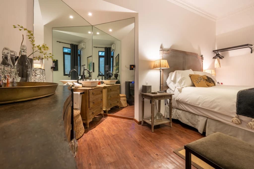 A room with wood flooring: a woven cane headboard and white bedding on one side, and an open bathroom with a wooden double vanity facing the mirror on the other