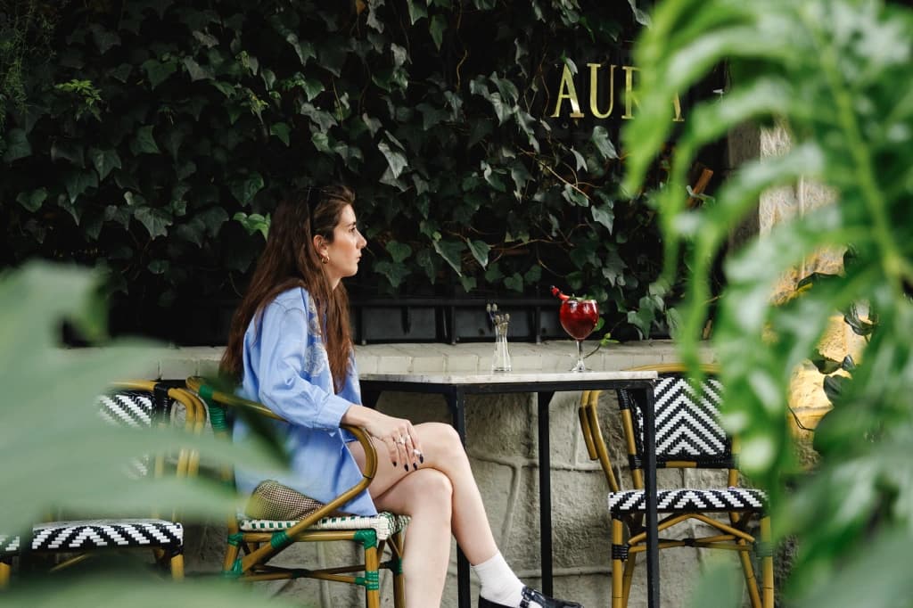A guest seated on Aura's terrace, a red cocktail on the table and the gold «Aura» sign among the ivy behind her