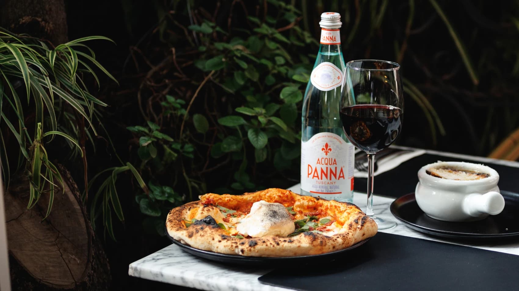 A wood-fired pizza with burrata on the terrace's marble table, beside a glass of red wine, Acqua Panna water and a bowl of gratinéed soup