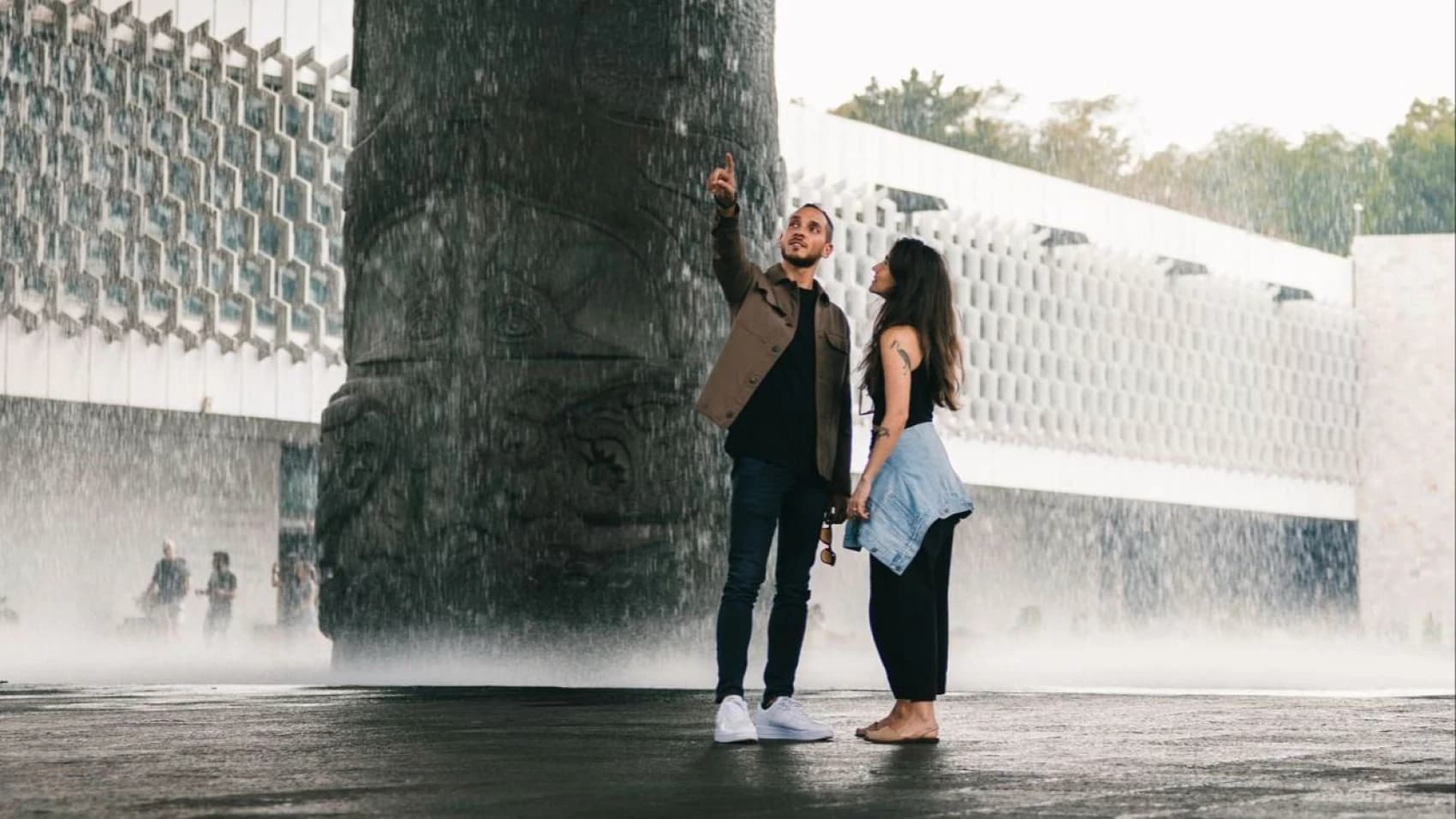 A couple beneath the water curtain of the National Anthropology Museum's Umbrella, him pointing at the reliefs on the central column