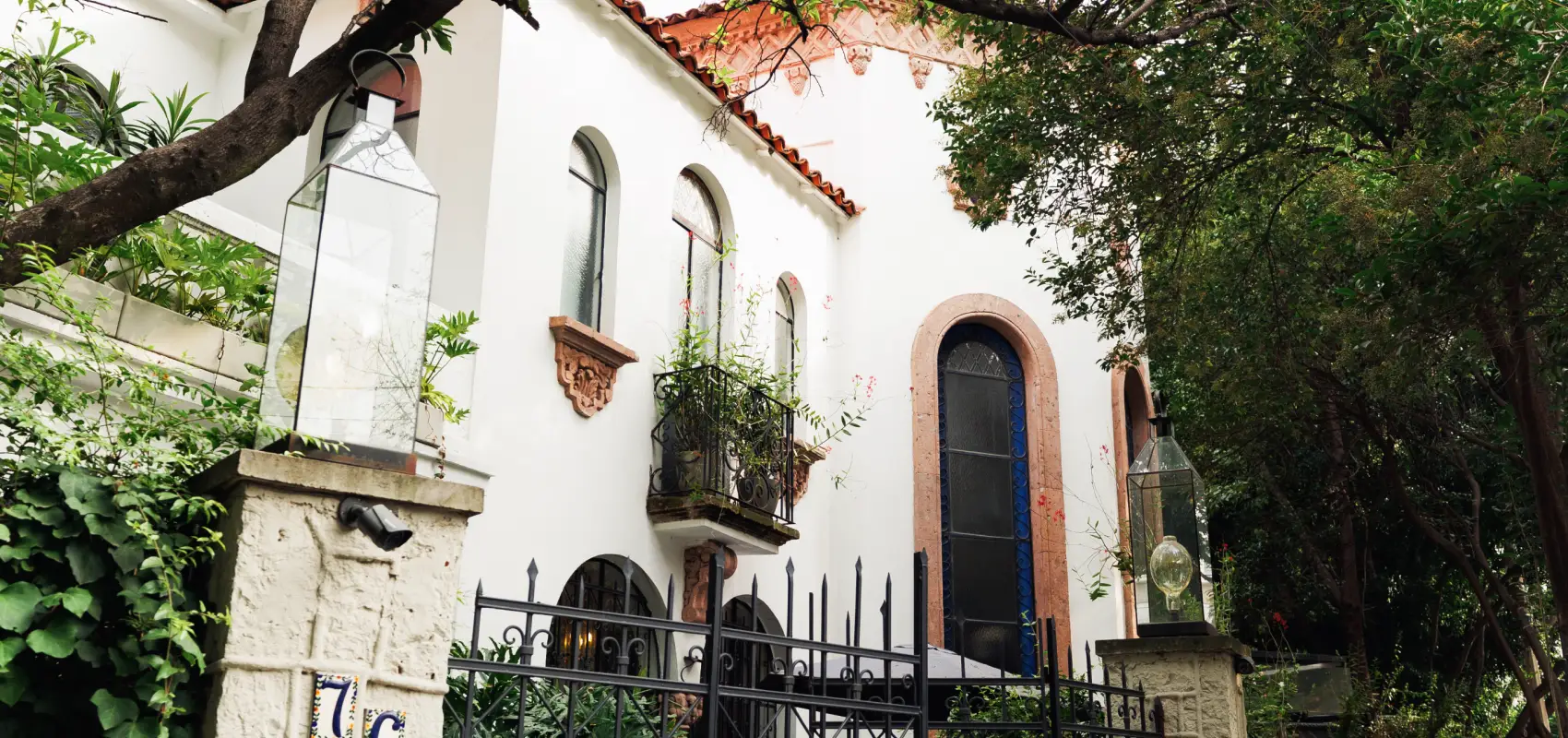 The white facade of the Polanco house among the trees, with a wrought-iron balcony, a blue door and the entrance gate